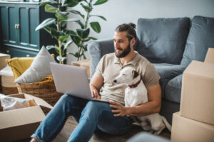 about-us-image Young men moving in new house. He is happy and using laptop to organize everything. His pet dog is with him.
